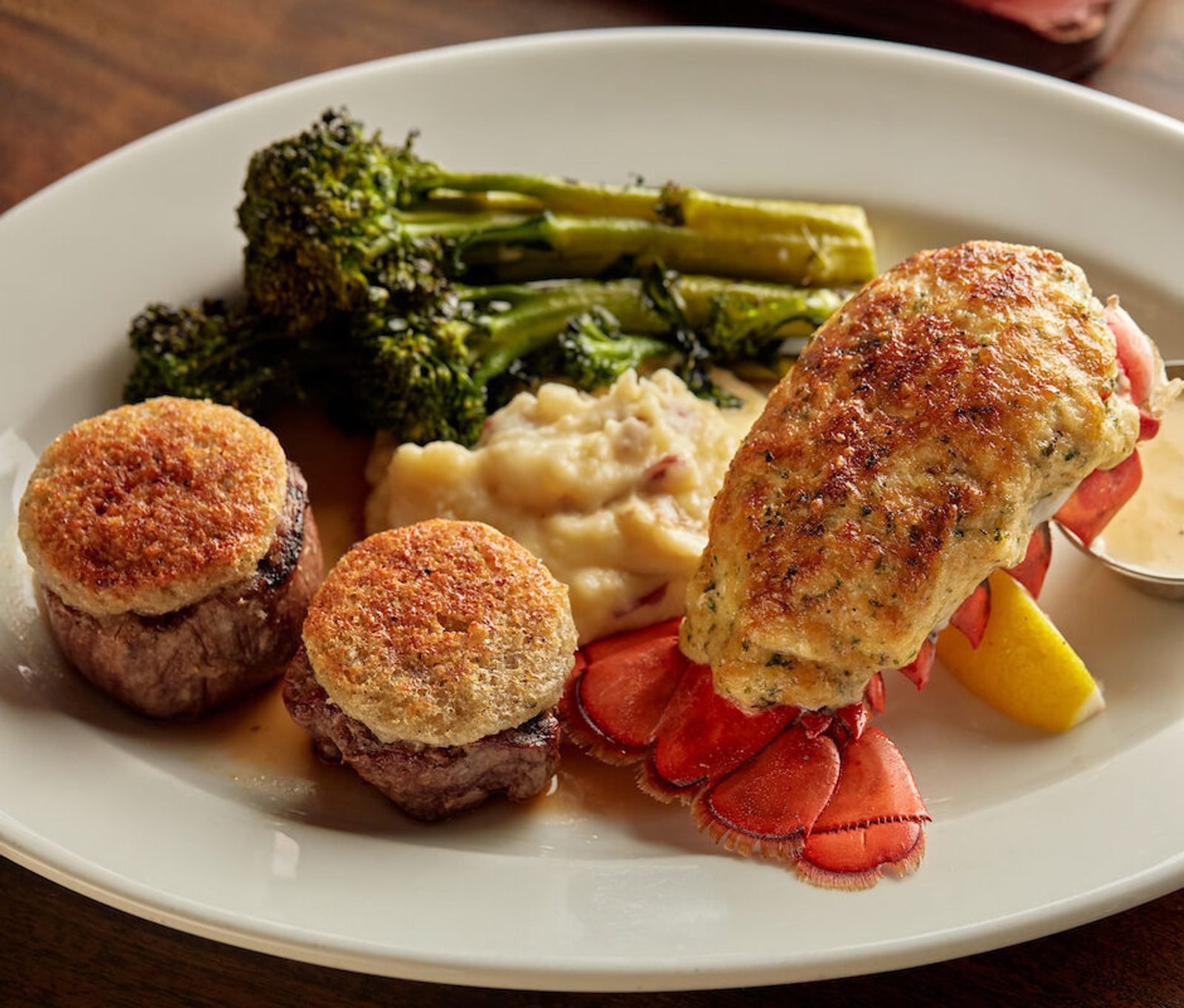 Plated surf and turf with two filet mignon medallions, lobster tail, roasted broccoli, mashed potatoes, and a lemon wedge on a white plate