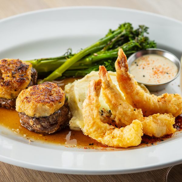 Steak medallions topped with crab cakes, tempura shrimp, mashed potatoes, broccolini, sauce, and a glass of wine.