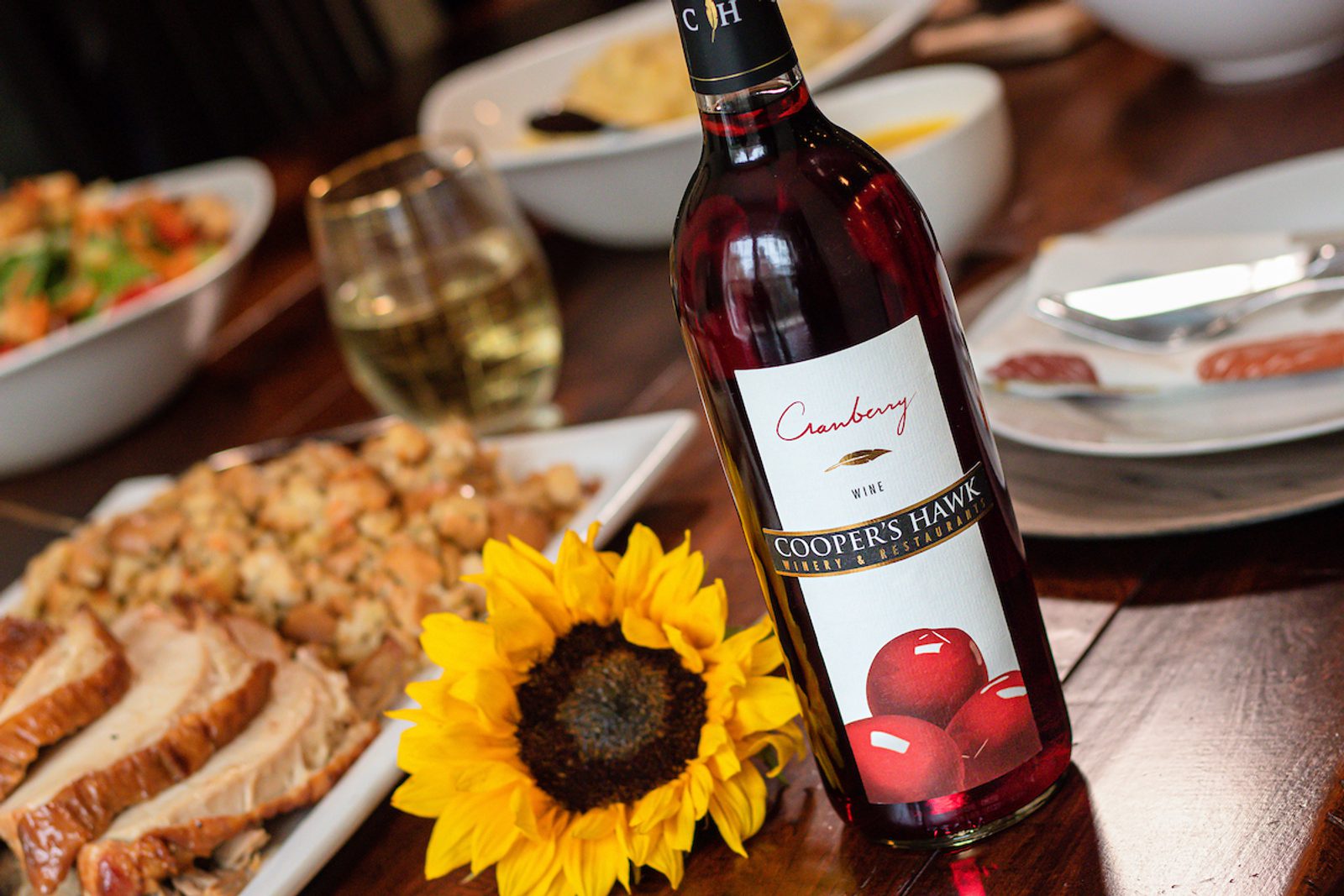 Bottle of cranberry wine on a dining table beside a sunflower, a glass of white wine, sliced meat, and serving dishes with food in the background