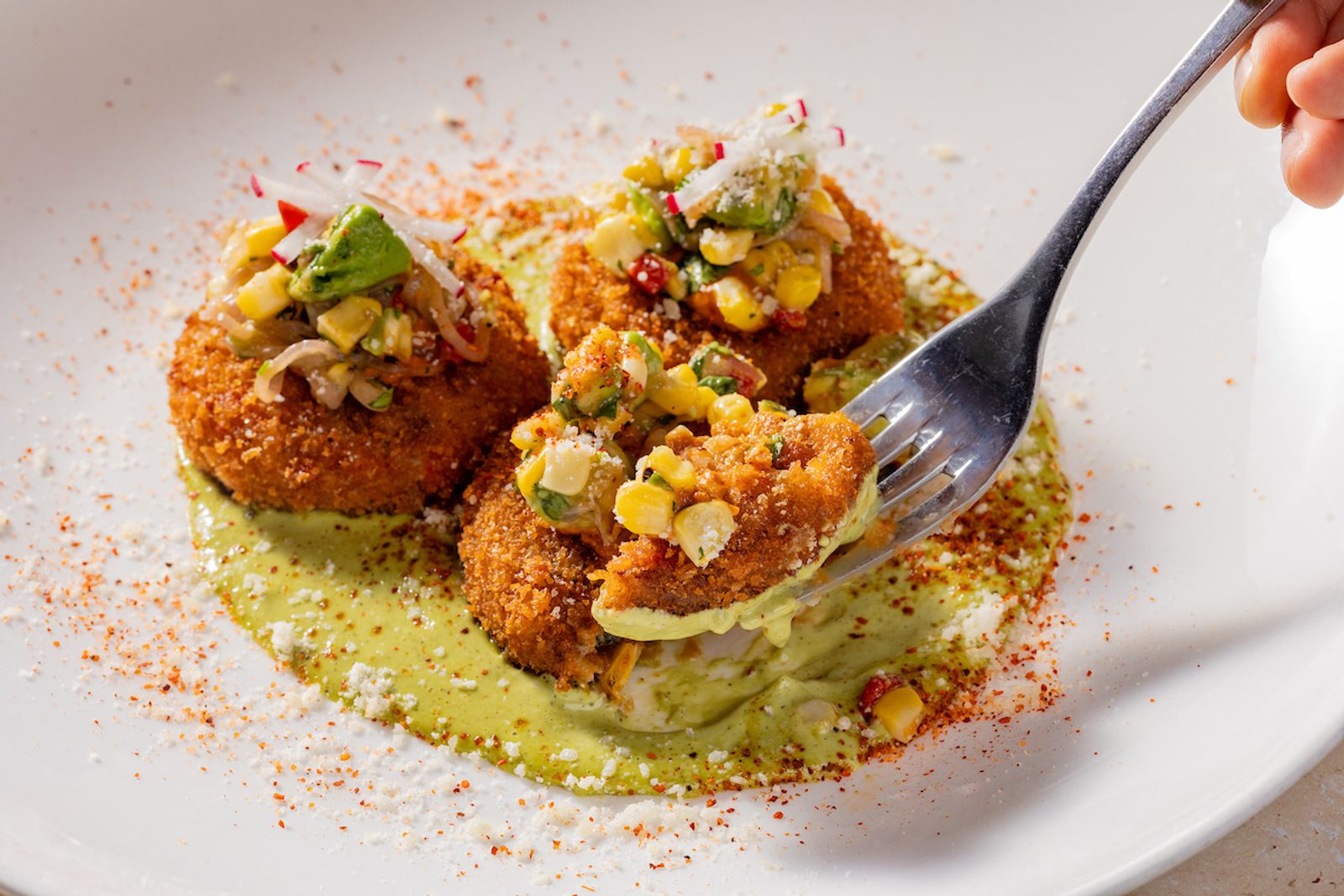 Three breaded croquettes topped with corn and avocado salsa on green sauce, a fork cutting one on a white plate