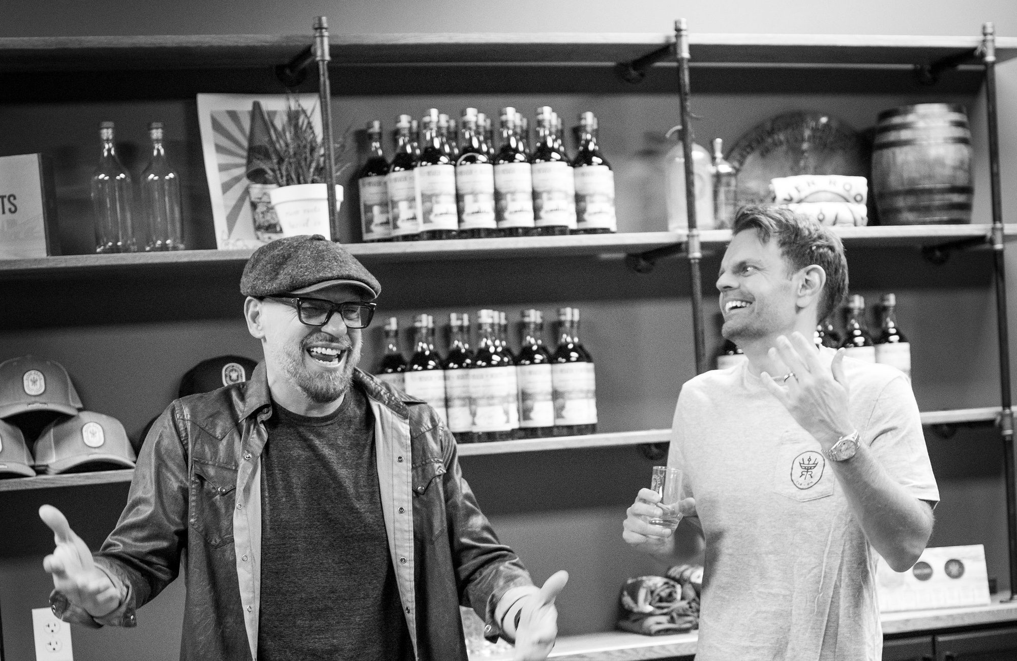 Two men laughing and holding tasting glasses in a room with rows of bottles on a table and shelves behind them