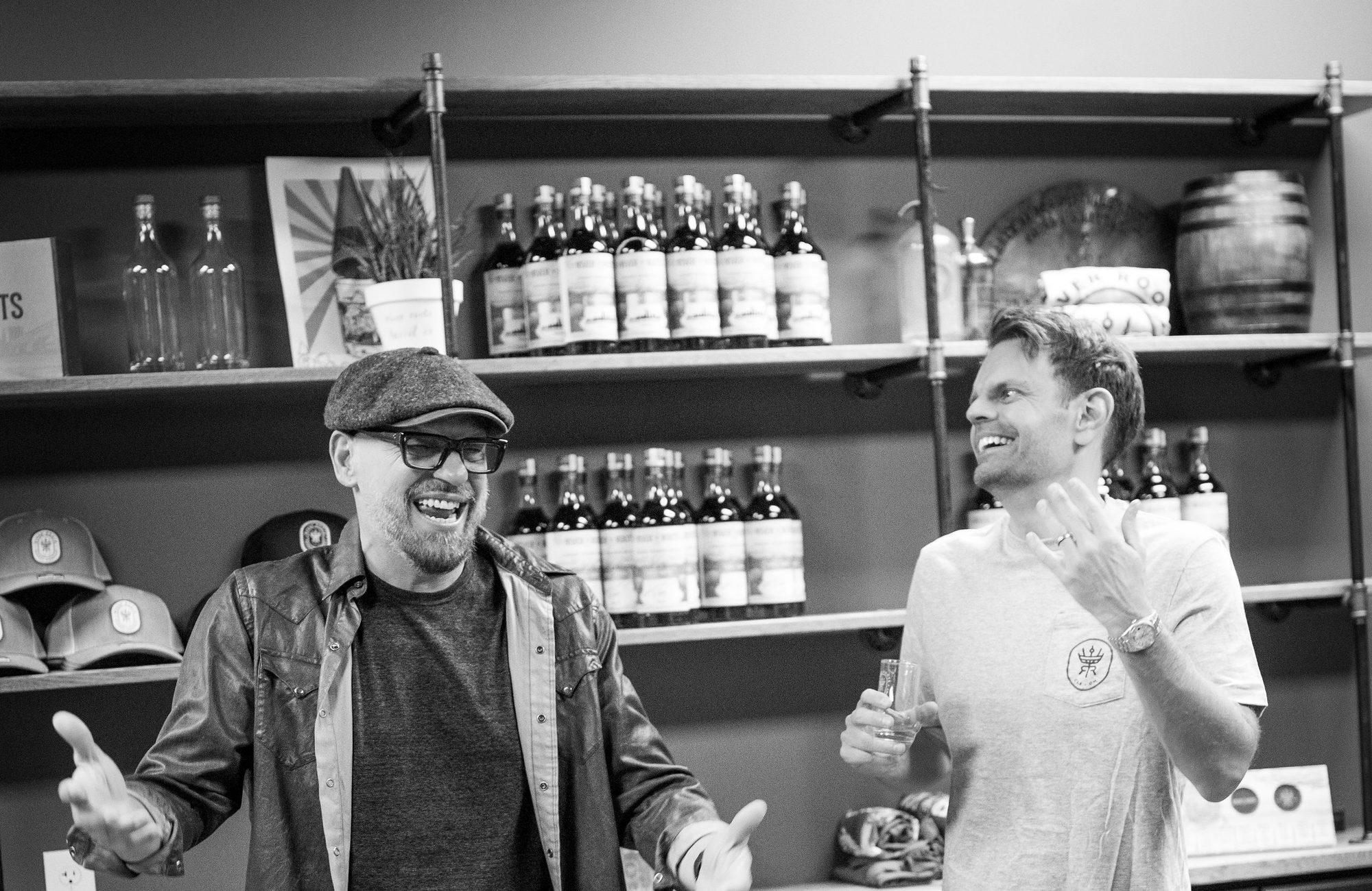 Two men laughing and holding tasting glasses in a room with rows of bottles on a table and shelves behind them