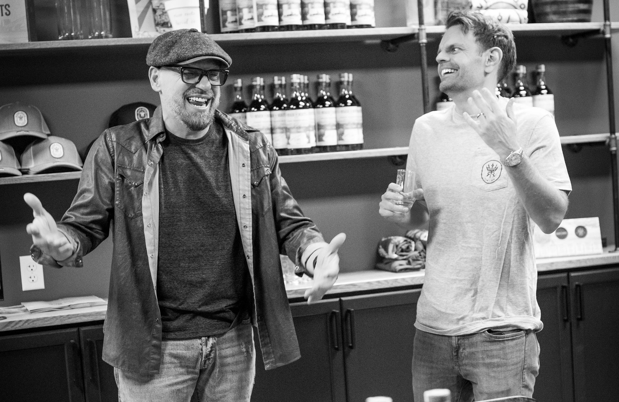Two men laughing and holding tasting glasses in a room with rows of bottles on a table and shelves behind them