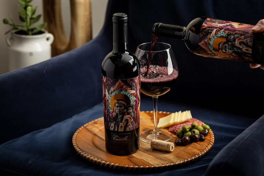 Red wine bottle with colorful illustrated label; wine being poured into glass on board with cheese, salami, olives and cork