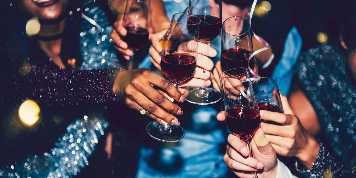 Group of friends smiling and toasting with red wine glasses at a lively party, with sparkling outfits and festive bokeh lights in the background