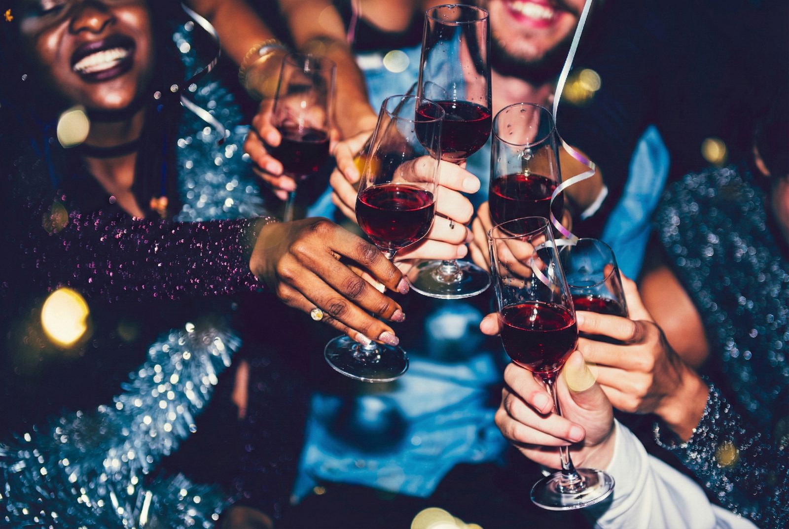Group of friends smiling and toasting with red wine glasses at a lively party, with sparkling outfits and festive bokeh lights in the background