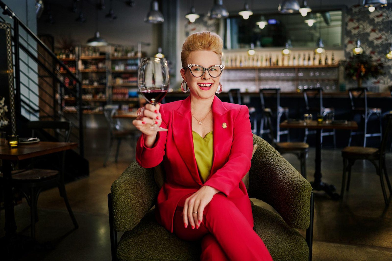 Person seated in a bar/lounge raising a glass of red wine toward the camera