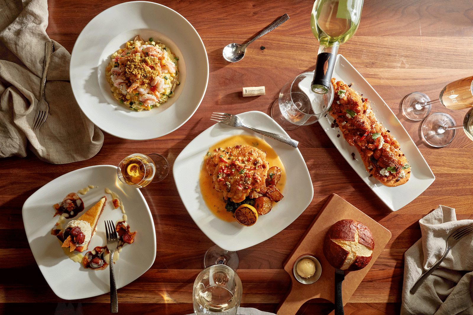 Overhead view of a restaurant table with several gourmet plated dishes, bread, dessert, wine glasses, and napkins on a warm wooden surface