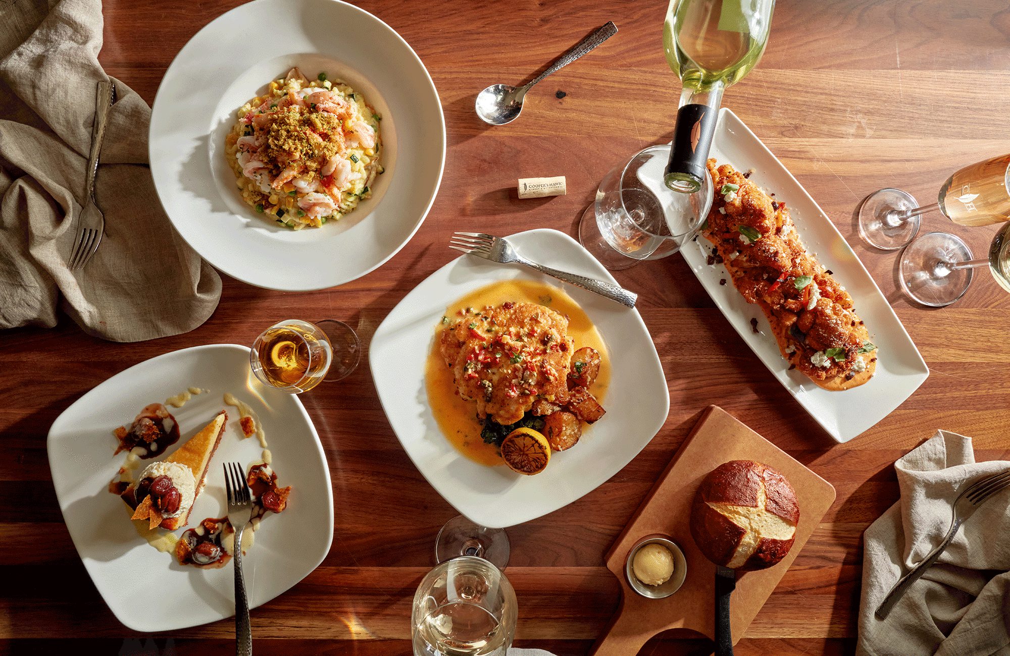 Overhead view of a restaurant table with several gourmet plated dishes, bread, dessert, wine glasses, and napkins on a warm wooden surface