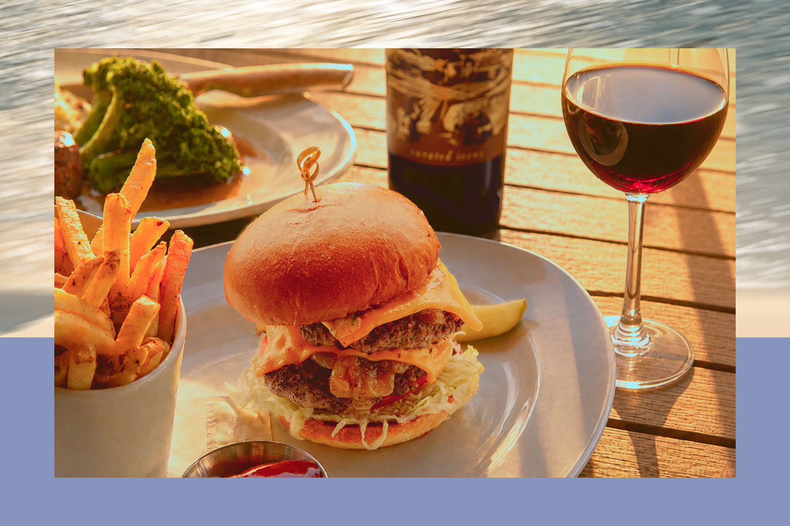 Double cheeseburger with lettuce and melted cheese, fries in a cup, and a glass of red wine on a wooden table