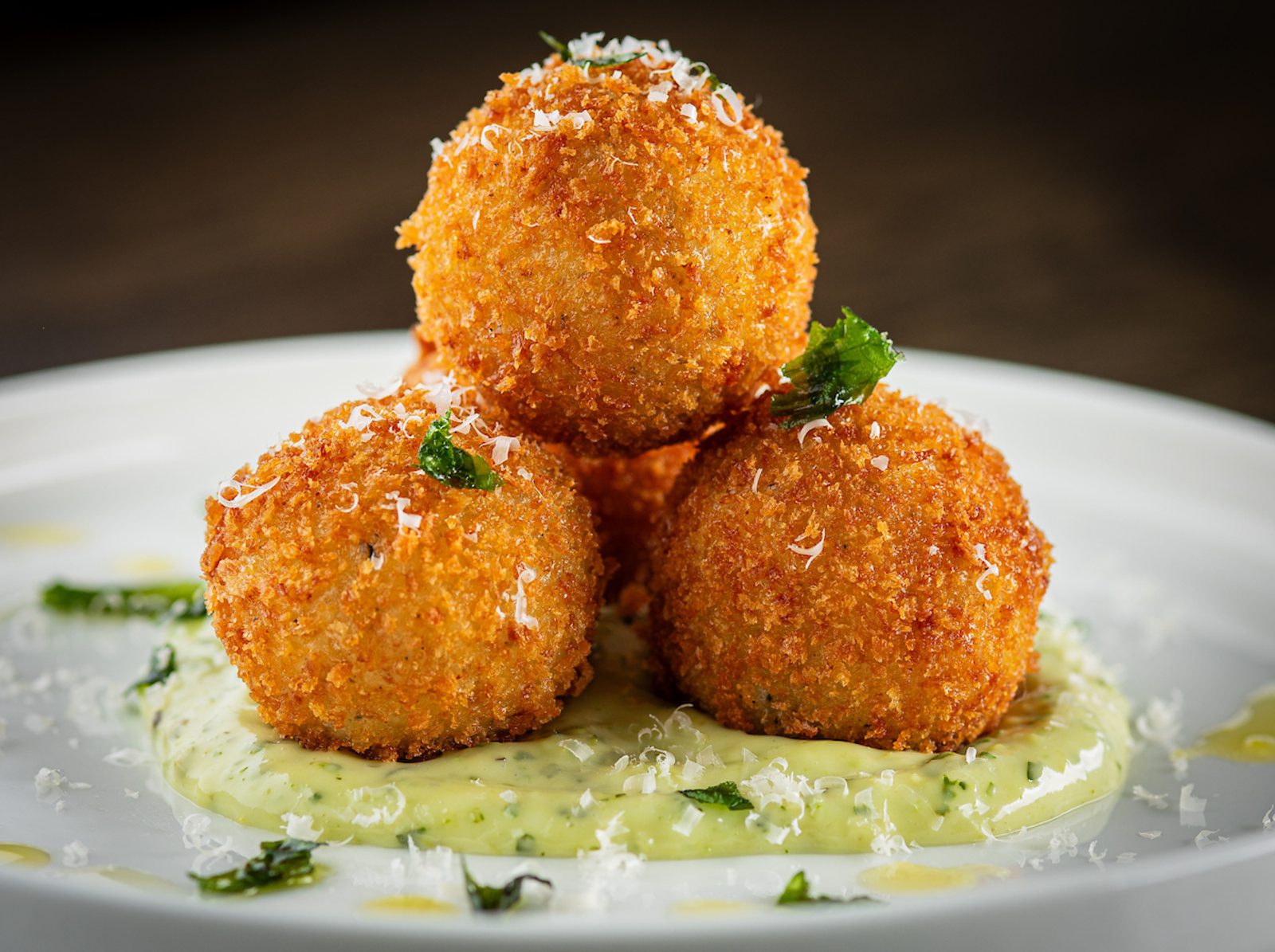Three crispy golden arancini balls stacked on a white plate over creamy green sauce, topped with herbs and grated cheese