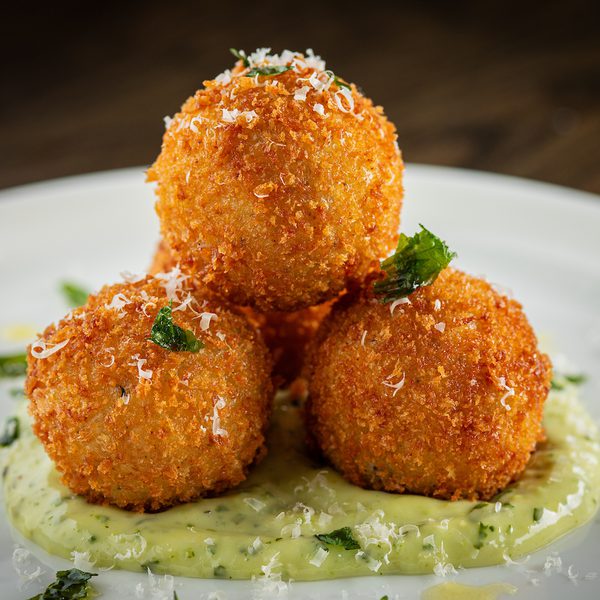 Three golden arancini (fried rice balls) stacked on green herb sauce, sprinkled with grated cheese and herbs