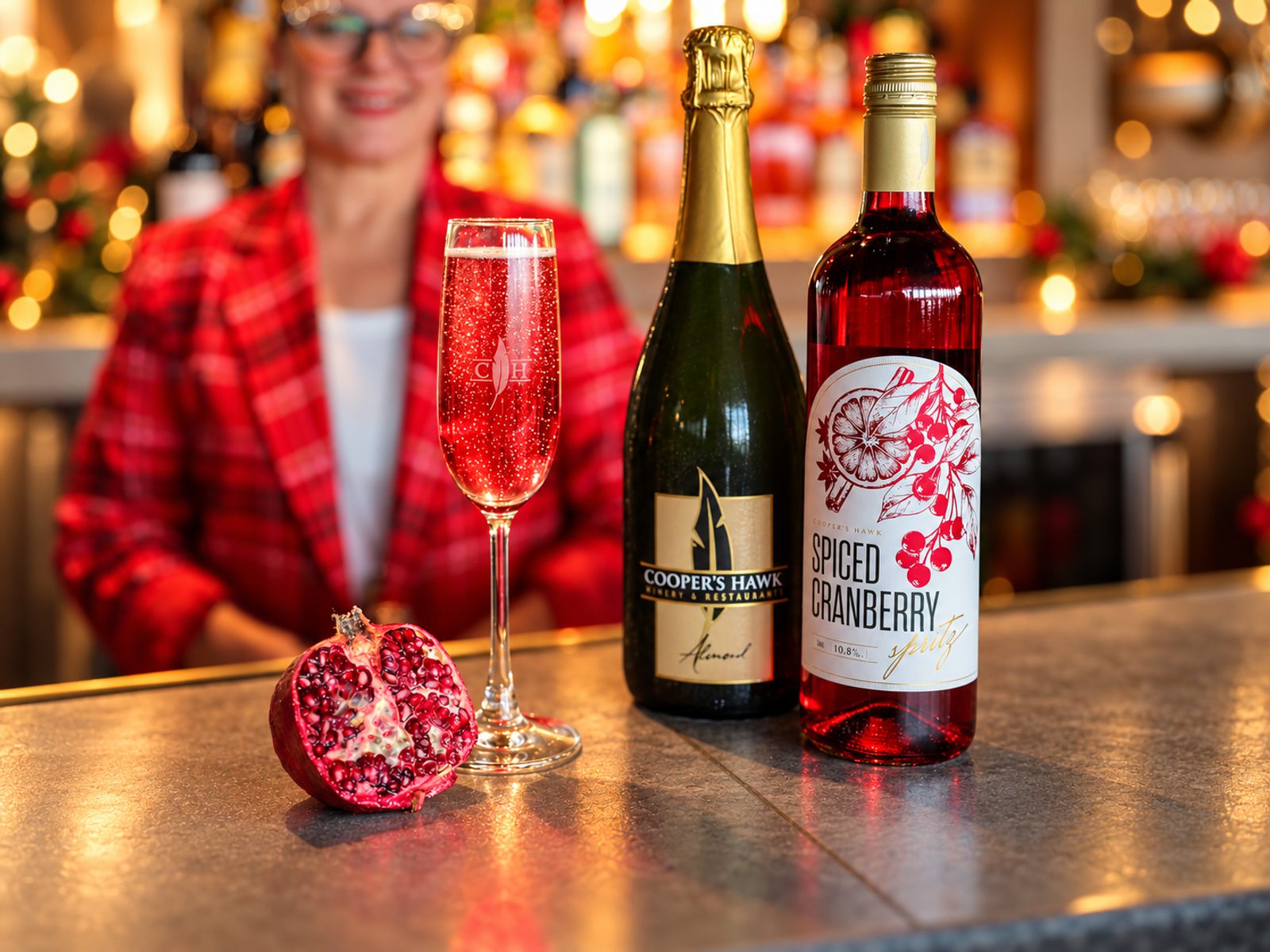 Festive bar scene with a blurred person in a red plaid jacket behind a glass of sparkling red drink, two wine bottles, and a halved pomegranate on the counter