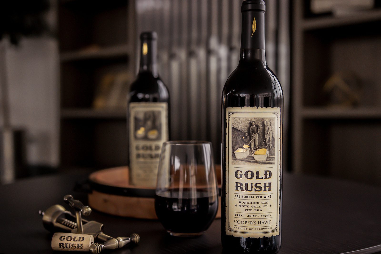 Bottle labeled GOLD RUSH and California Red Wine next to a filled wine glass and corkscrew; second bottle blurred