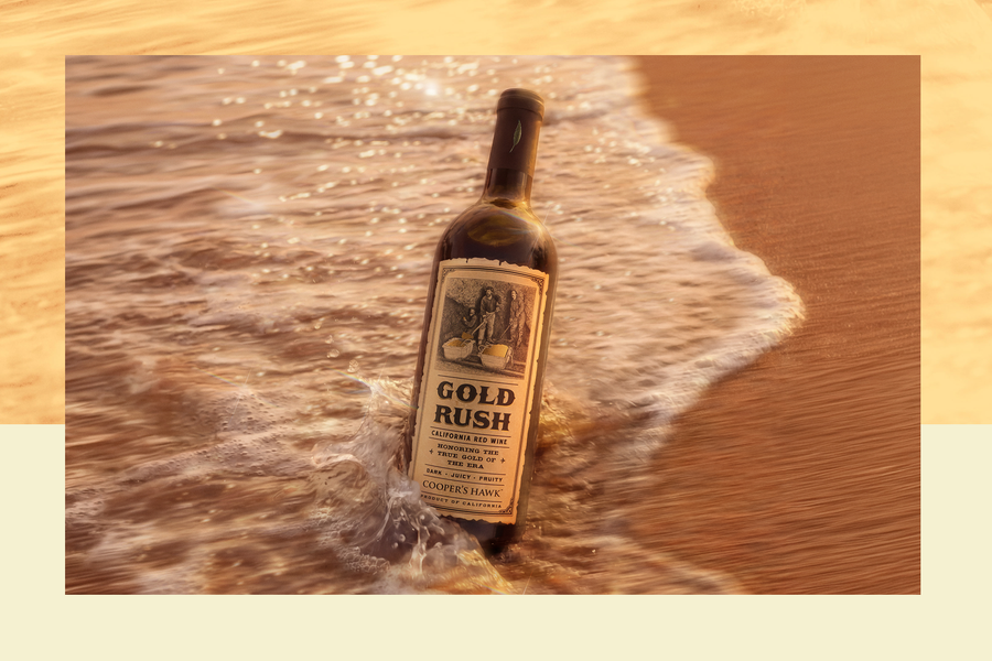Bottle of wine labeled GOLD RUSH California Red Wine Cooper's Hawk, partially in surf on sandy shore at sunset