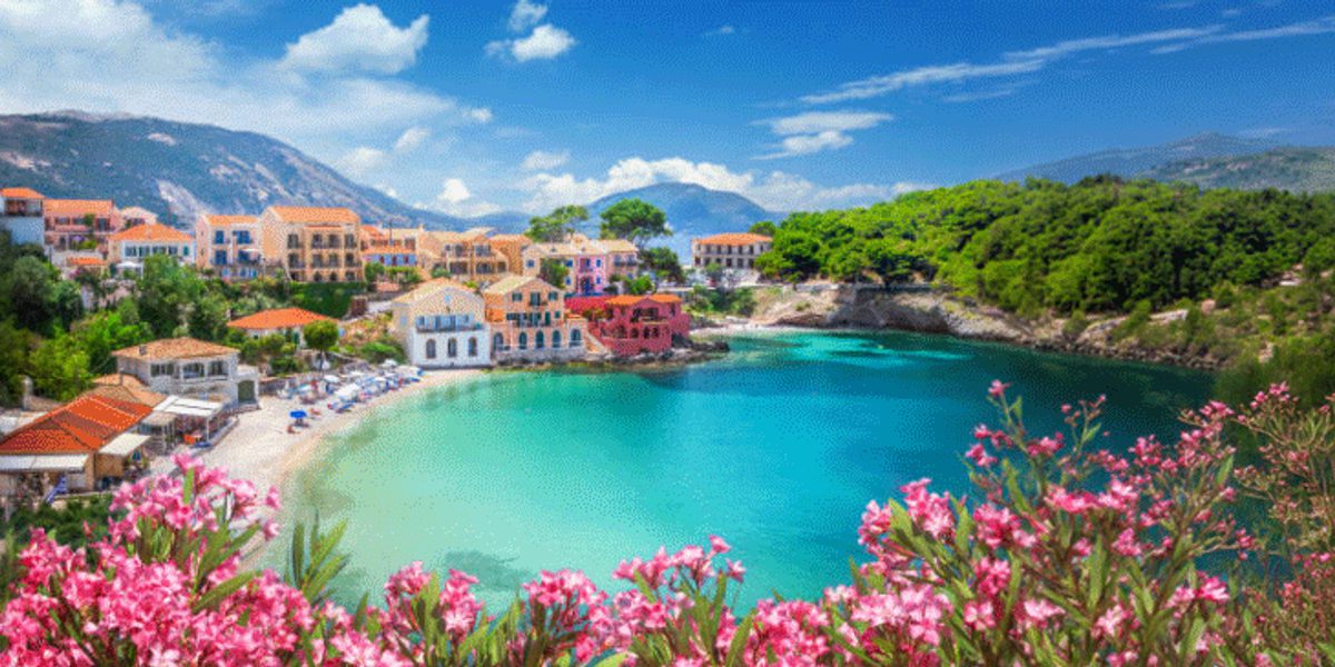 Panoramic coastal cove with turquoise water, small colorful seaside village, pink flowering shrubs and forested hills.