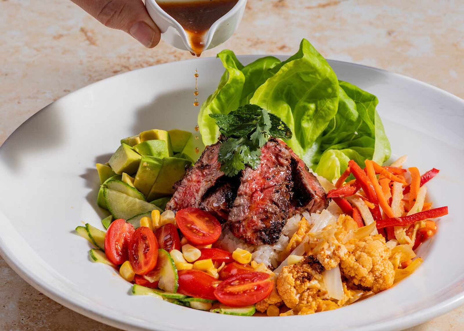 Rice bowl with sliced steak, avocado, lettuce, cherry tomatoes, corn, cauliflower and carrots; dressing being poured.