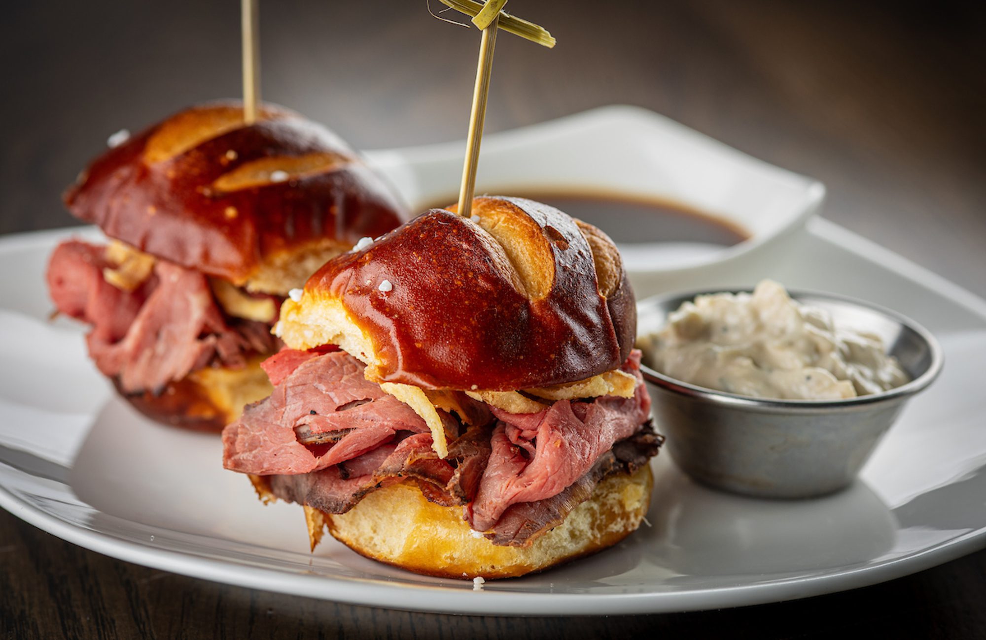 Two roast beef sliders on pretzel buns with skewers, served with a cup of au jus and a ramekin of horseradish sauce