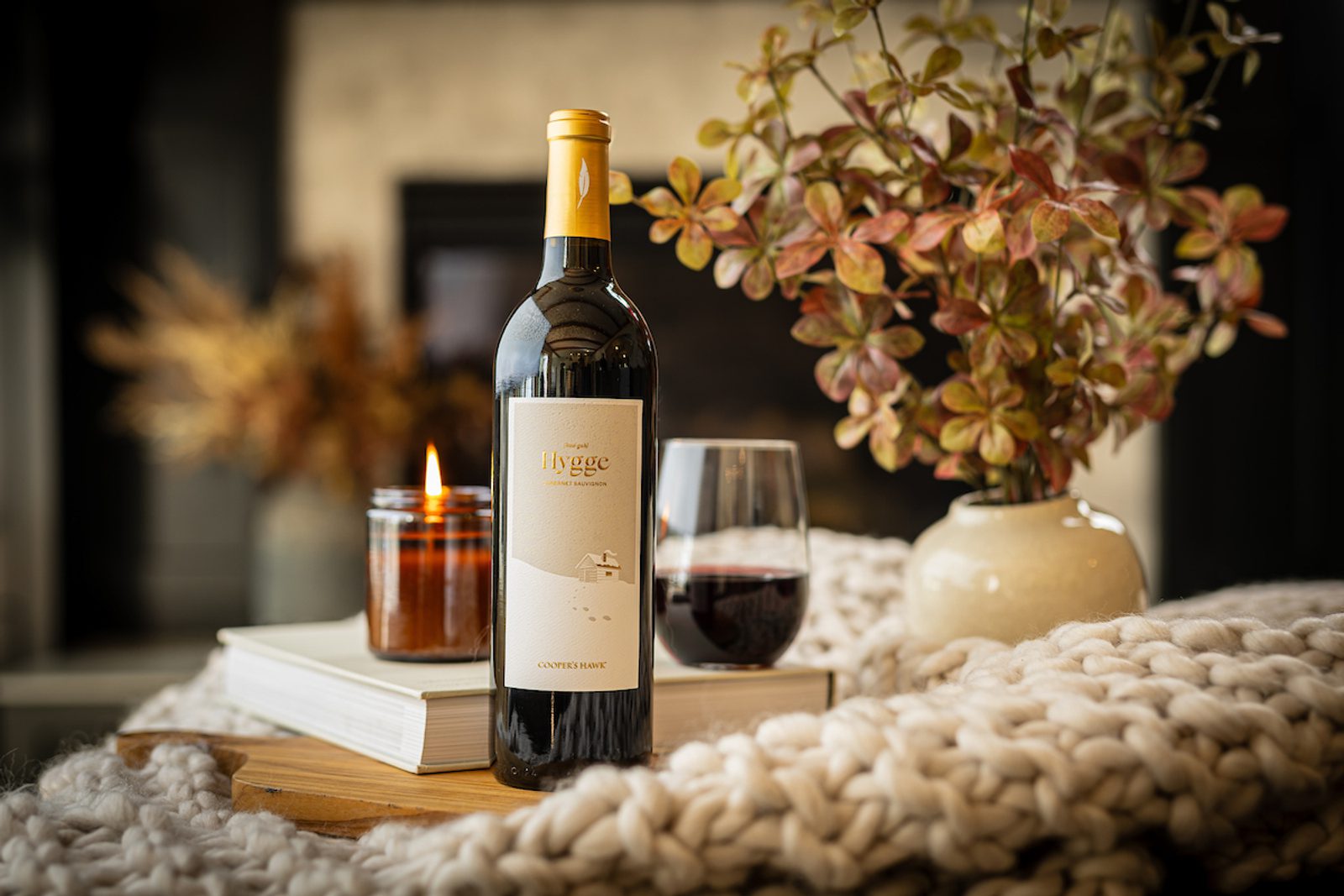 Bottle labeled Hygge Cabernet Sauvignon Coopers Hawk next to a lit candle, glass of red wine, book and vase on chunky knit blanket