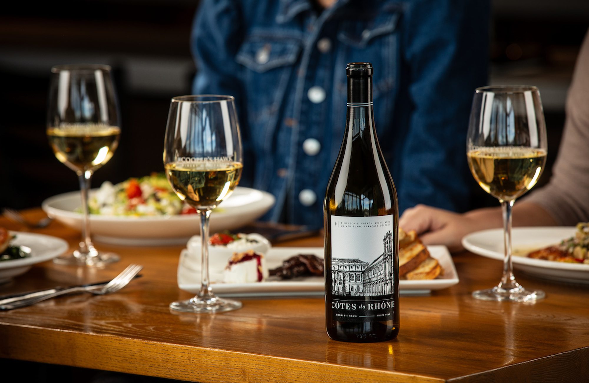 Wine bottle labeled Côtes du Rhône on a wooden table with three glasses of white wine and plates of food