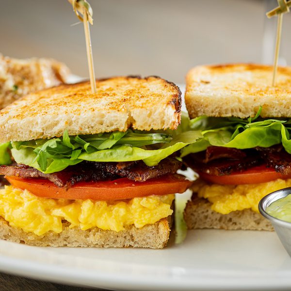 Breakfast sandwich with scrambled eggs, tomato, bacon and lettuce on toasted bread, served with green dipping sauce.