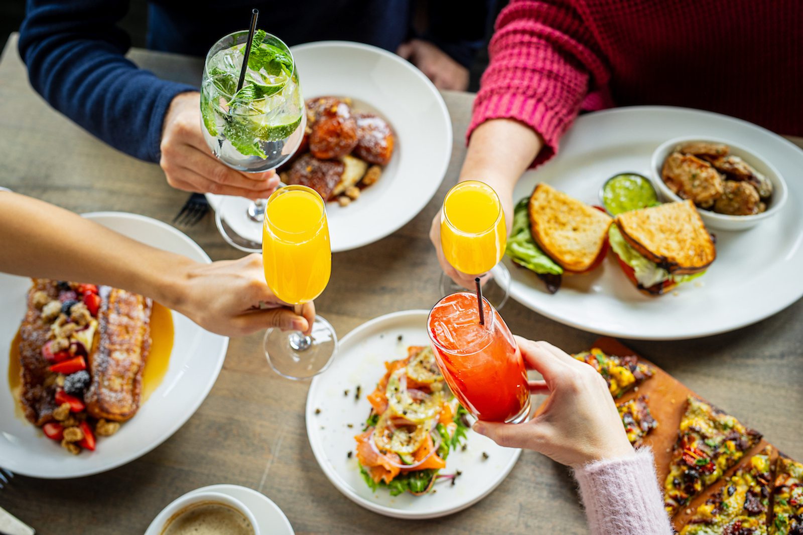 Hands raising colorful drinks over a table of brunch dishes, including sandwiches, toast, French toast, salads, and coffee in a lively dining setting