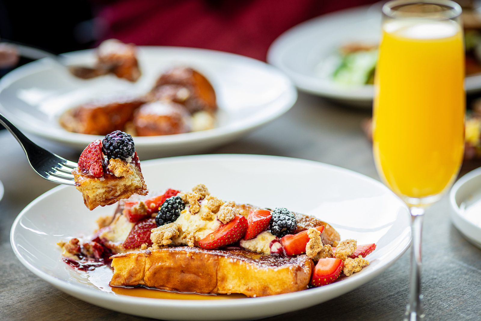 French toast topped with strawberries, blackberries, and crumb topping on a white plate, with a fork holding a bite and a glass of orange juice nearby