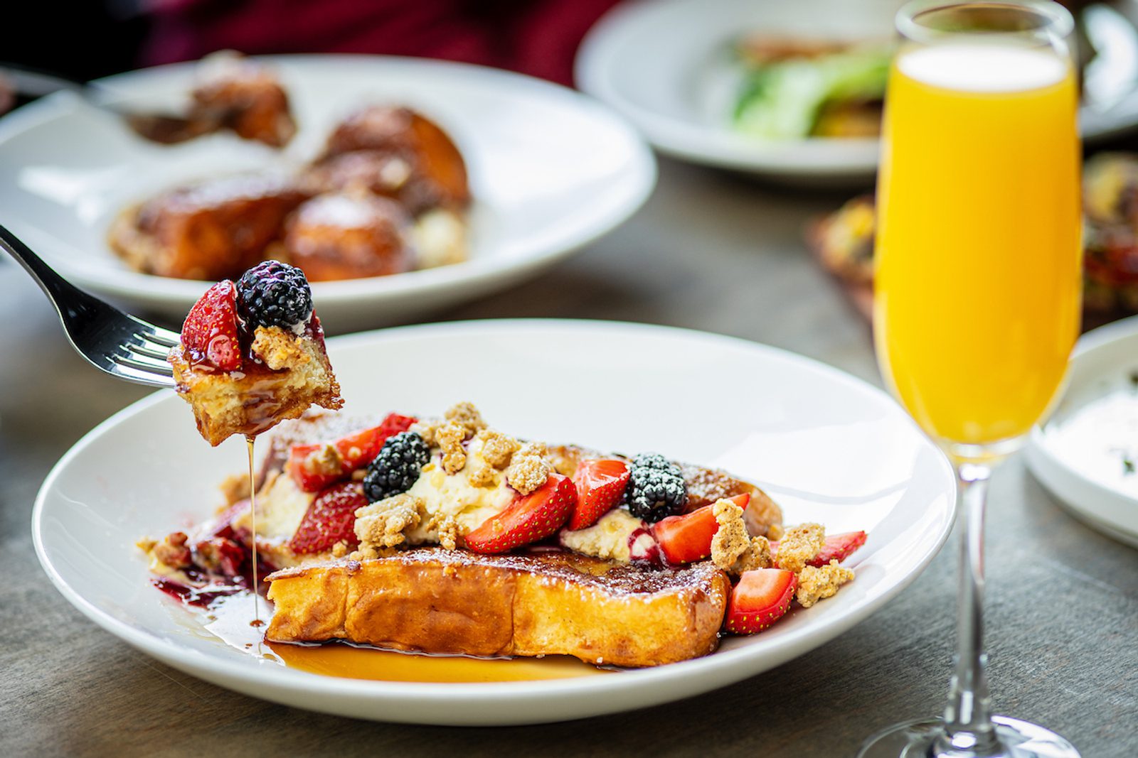 French toast topped with strawberries, blackberries, crumbled streusel and syrup, with a glass of orange beverage.