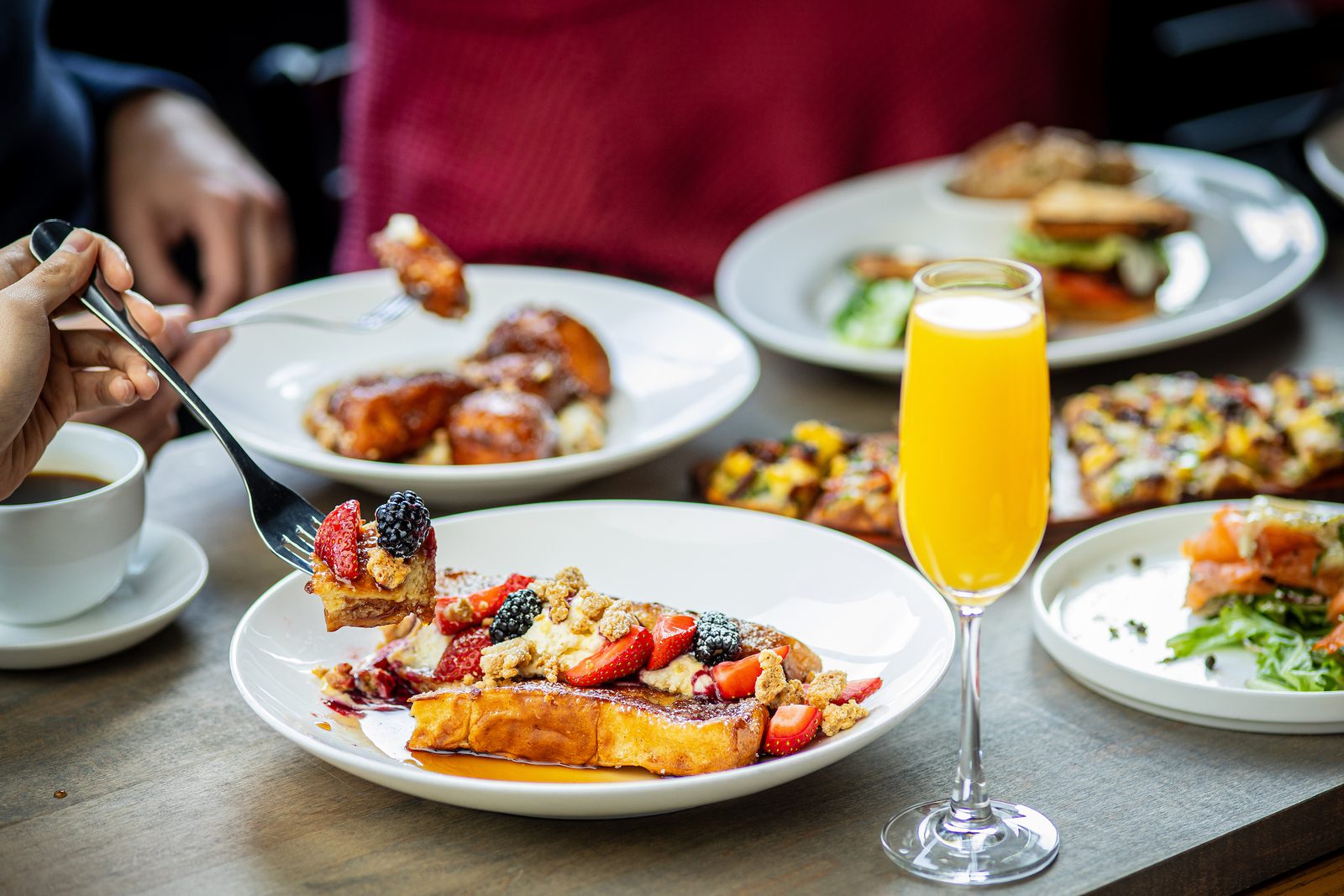 A hand lifts a fork with a piece of French toast topped with berries, a glass of mimosa nearby, and various dishes on the table.