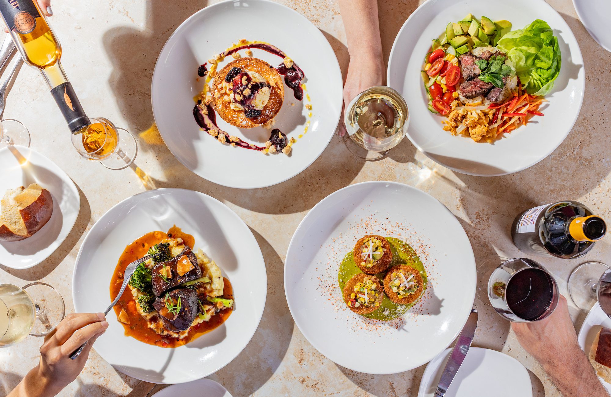 Overhead table with multiple plated entrees and desserts, wine glasses and hands pouring and holding wine