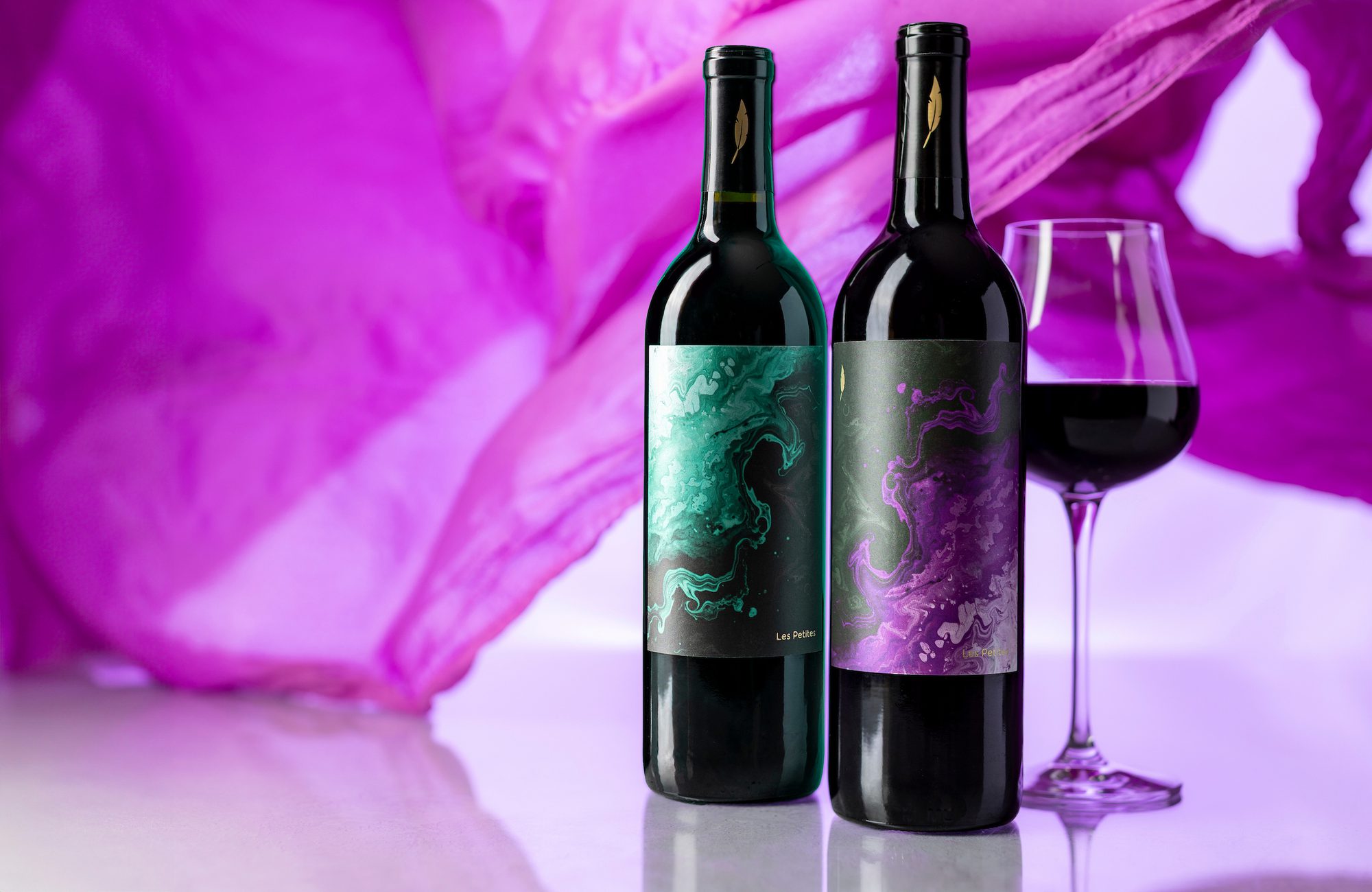 Two wine bottles with green and purple marbled labels and a glass of red wine against a purple fabric backdrop