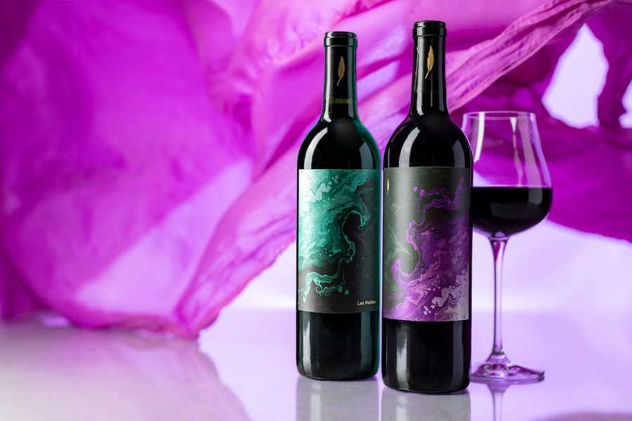 Two red-wine bottles with abstract green and purple labels next to a filled wine glass, purple fabric backdrop