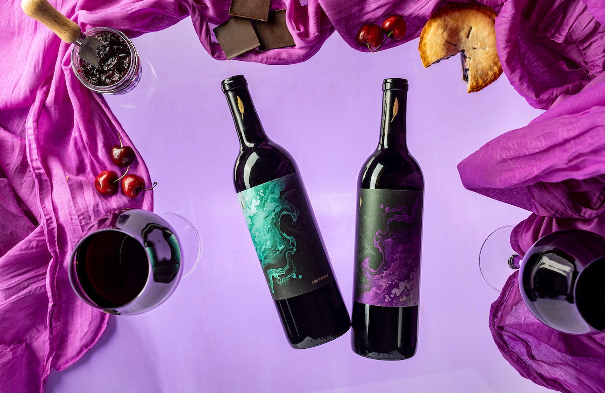 Two wine bottles with teal and purple marbled labels surrounded by wine glasses, cherries, jam, chocolate, and pie on purple cloth