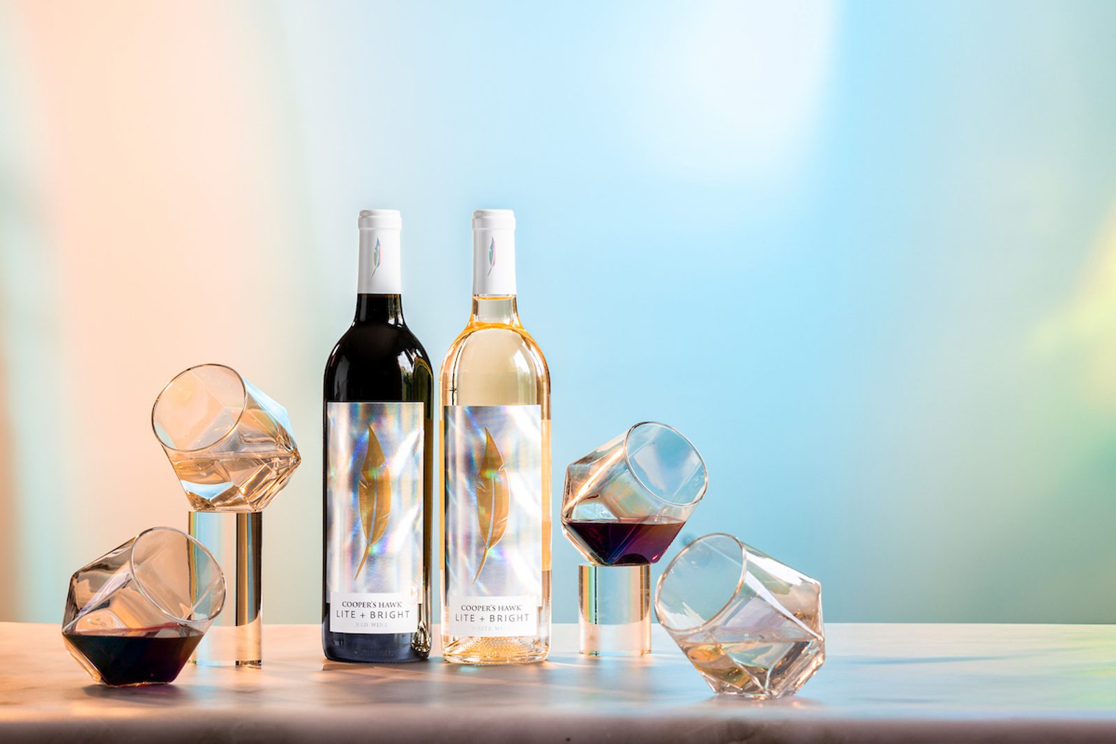 Two wine bottles labeled Cooper's Hawk Lite + Bright (red and white) with tilted wine glasses on metal stands