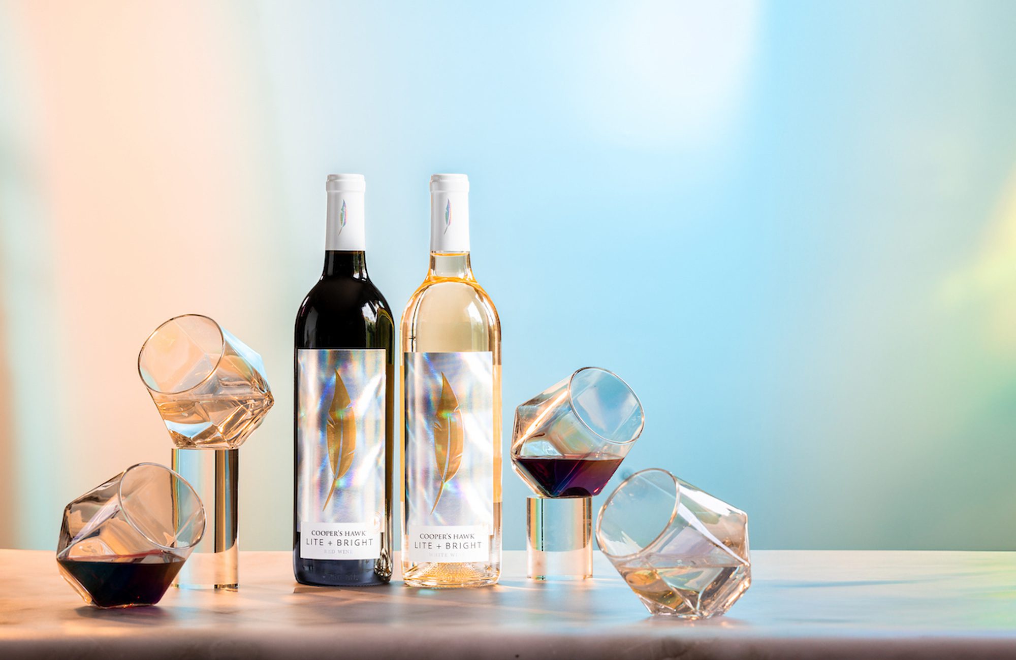 Two wine bottles labeled Cooper's Hawk Lite + Bright (red and white) with tilted wine glasses on metal stands