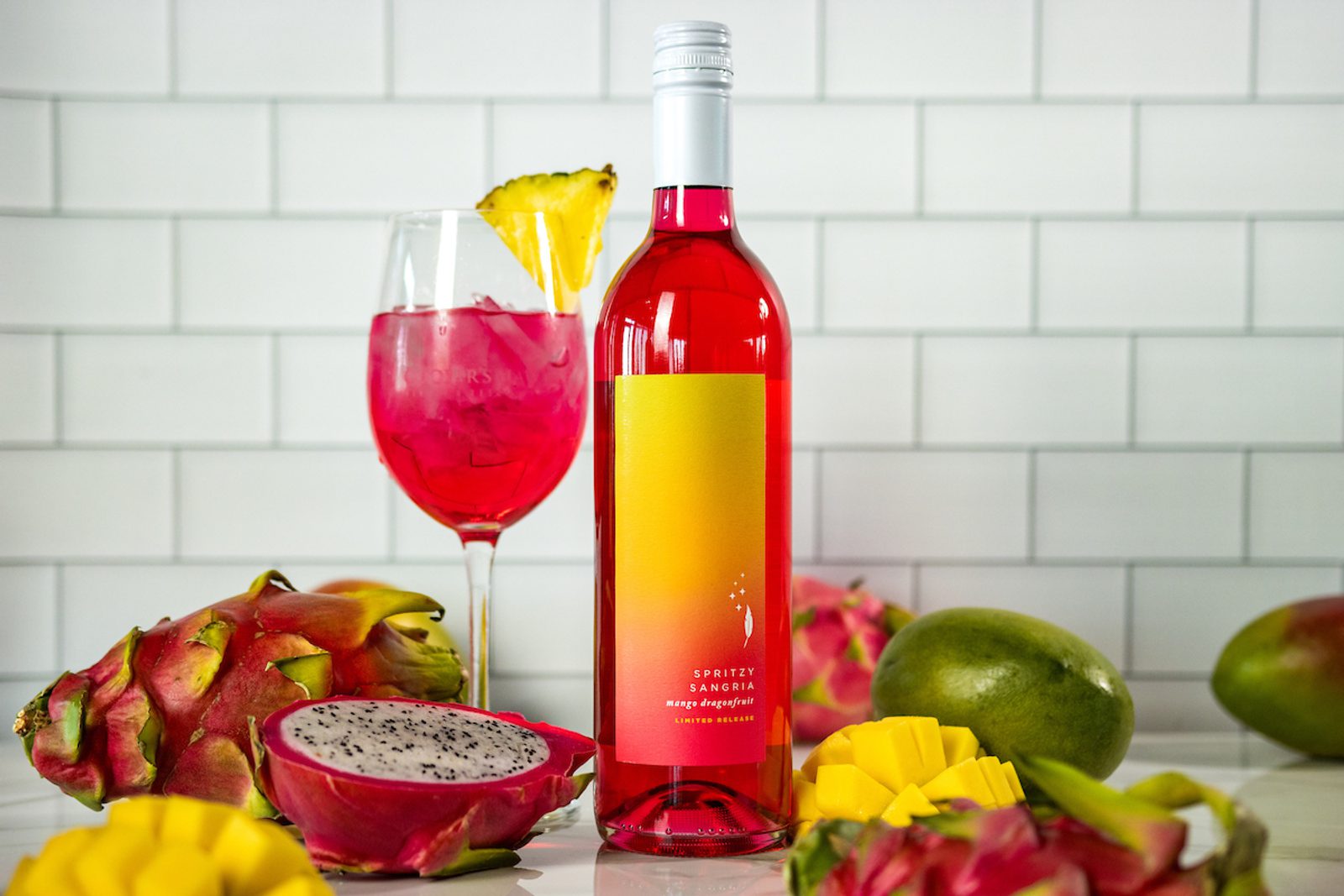 Bottle labeled Spritzy Sangria mango dragonfruit beside a glass of pink sangria with pineapple wedge, dragonfruit and mango.