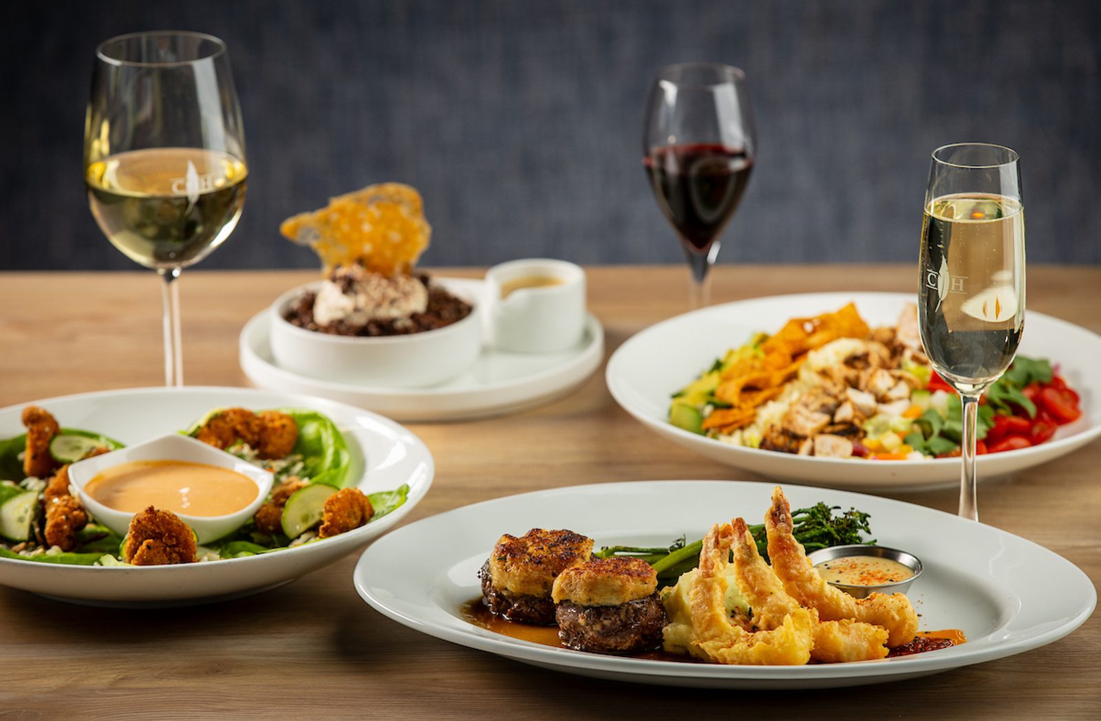 Restaurant table with plated dishes: beef medallions topped with crab cakes, tempura shrimp, a fried-shrimp salad, chopped salad, dessert and wine.