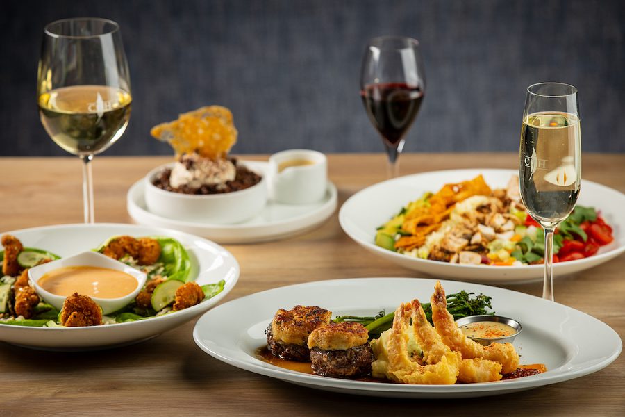 Restaurant table with plated dishes: beef medallions topped with crab cakes, tempura shrimp, a fried-shrimp salad, chopped salad, dessert and wine.