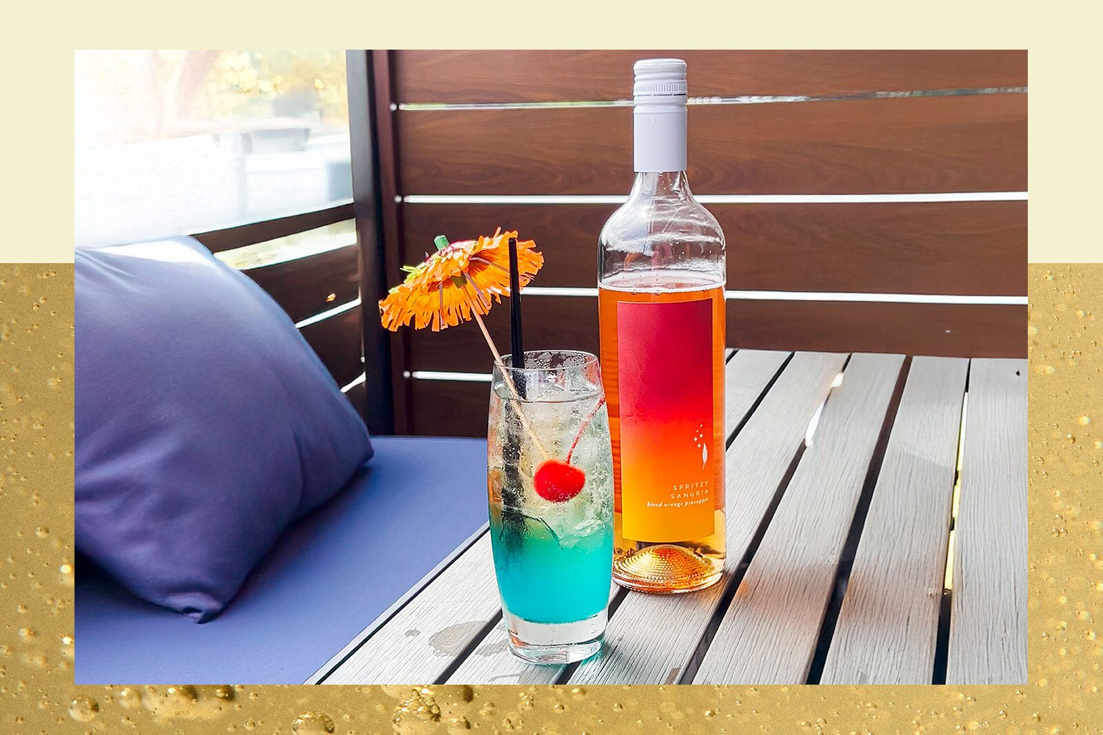 Bottle of spritz sangria and blue cocktail with cherry and orange paper umbrella on wood patio table.