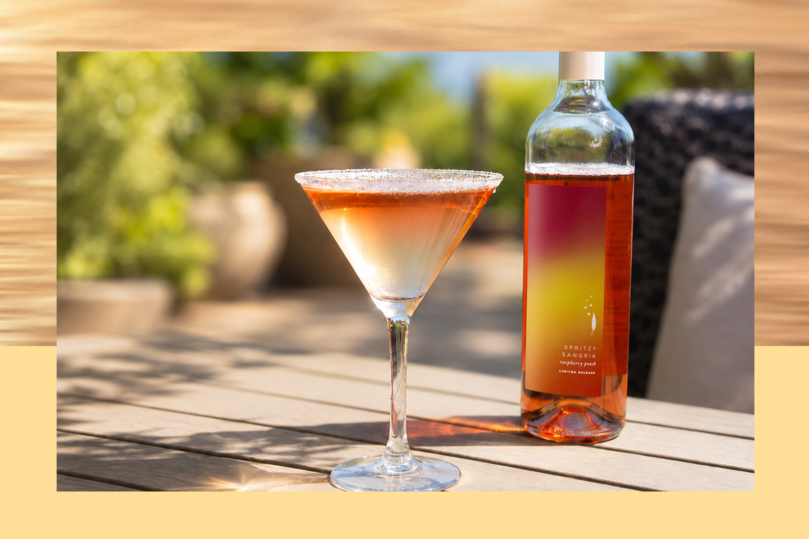Martini glass with amber-over-clear cocktail rimmed with sugar on a patio table beside a bottle labeled SPRITZY SANGRIA