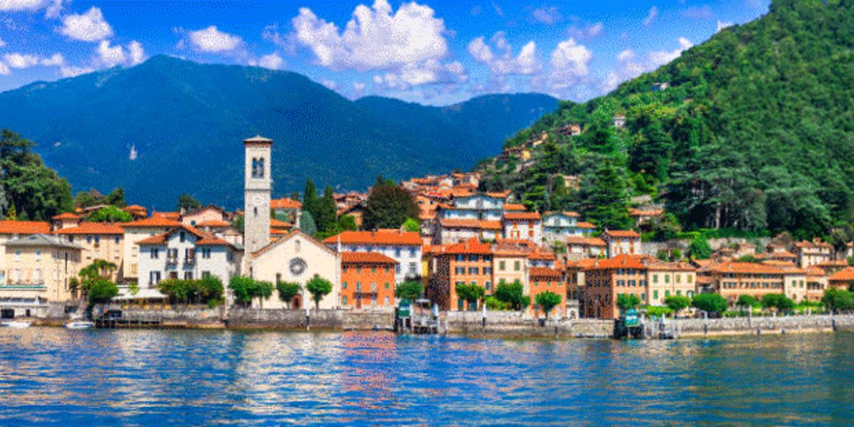Lakeside town with red-tiled roofs and church bell tower along a waterfront, backed by forested hills and distant mountains.