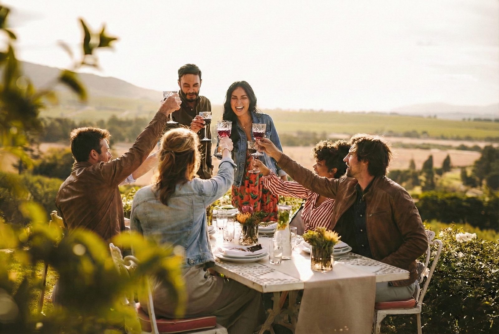 A group of friends toast with wine glasses around an outdoor table at sunset, smiling in a vineyard with hills and fields in the background.