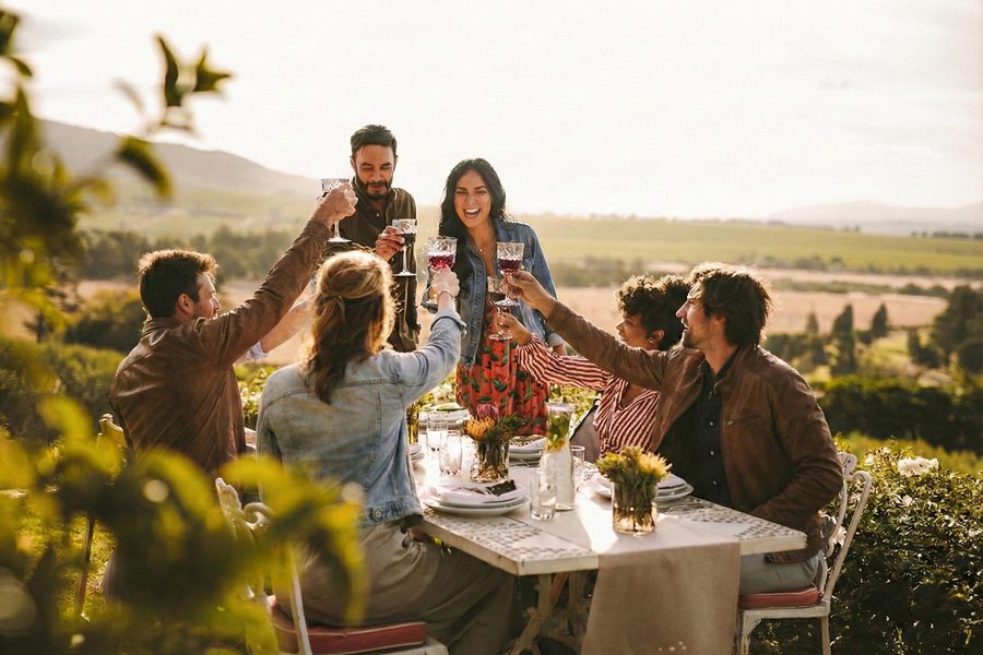 A group of friends toast with wine glasses around an outdoor table at sunset, smiling in a vineyard with hills and fields in the background.