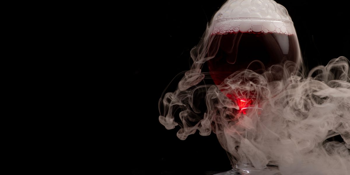 A glass goblet filled with dark red liquid, topped with foam and surrounded by swirling white smoke against a black background