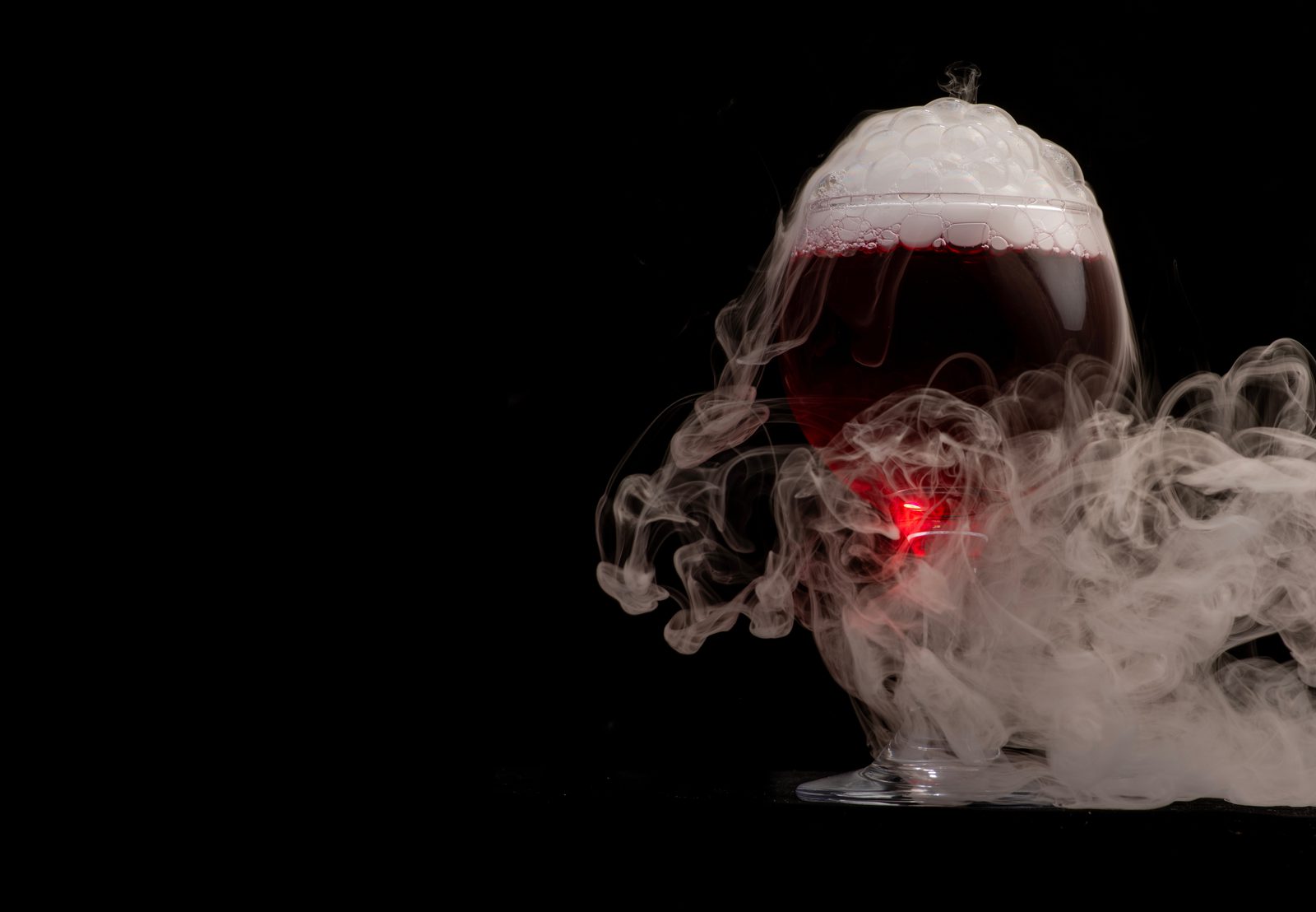 A glass goblet filled with dark red liquid, topped with foam and surrounded by swirling white smoke against a black background