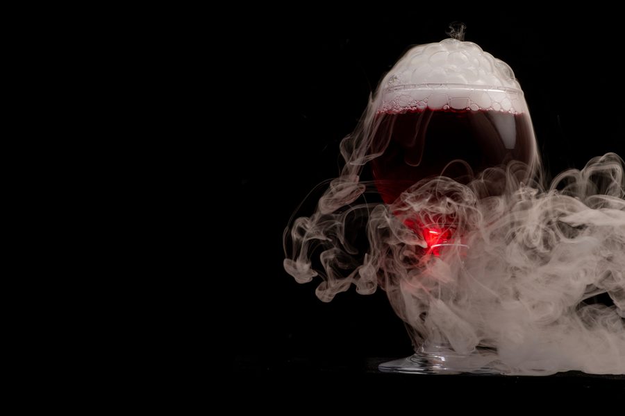 A glass goblet filled with dark red liquid, topped with foam and surrounded by swirling white smoke against a black background
