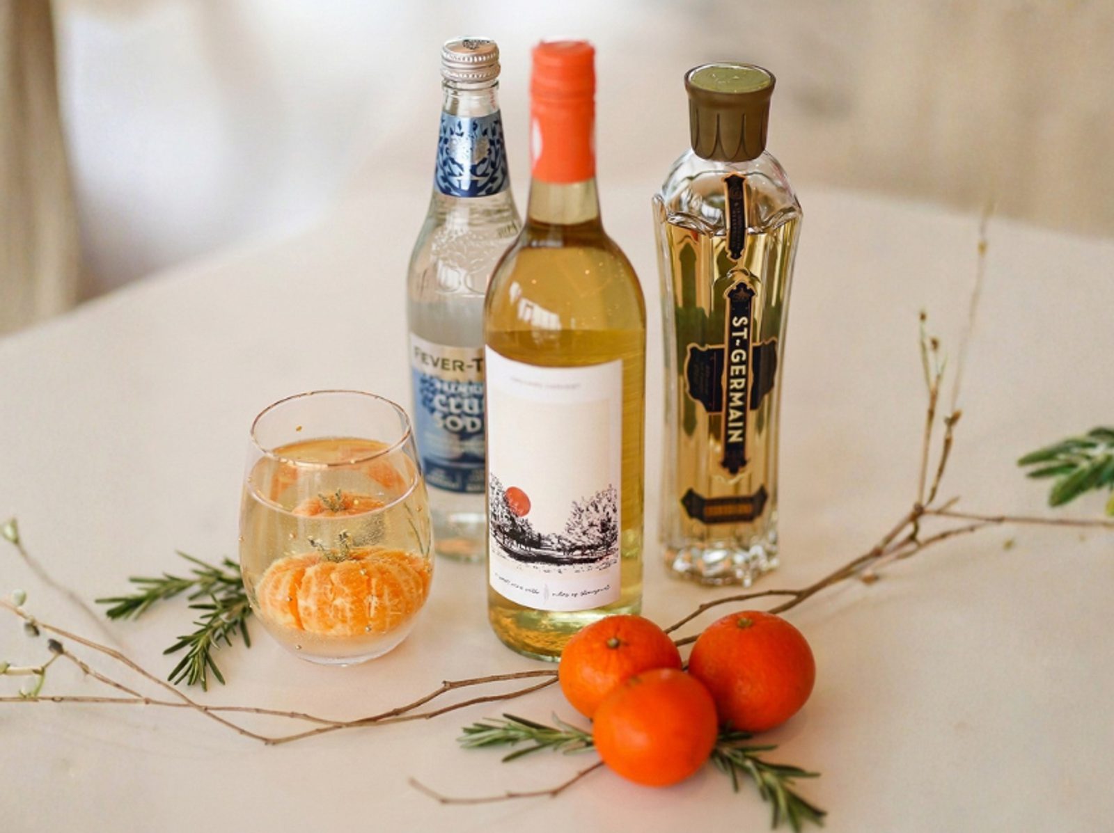 Three bottles of drink mixers and liqueur on a light table beside a glass with orange slices, tangerines, and sprigs of rosemary and twigs