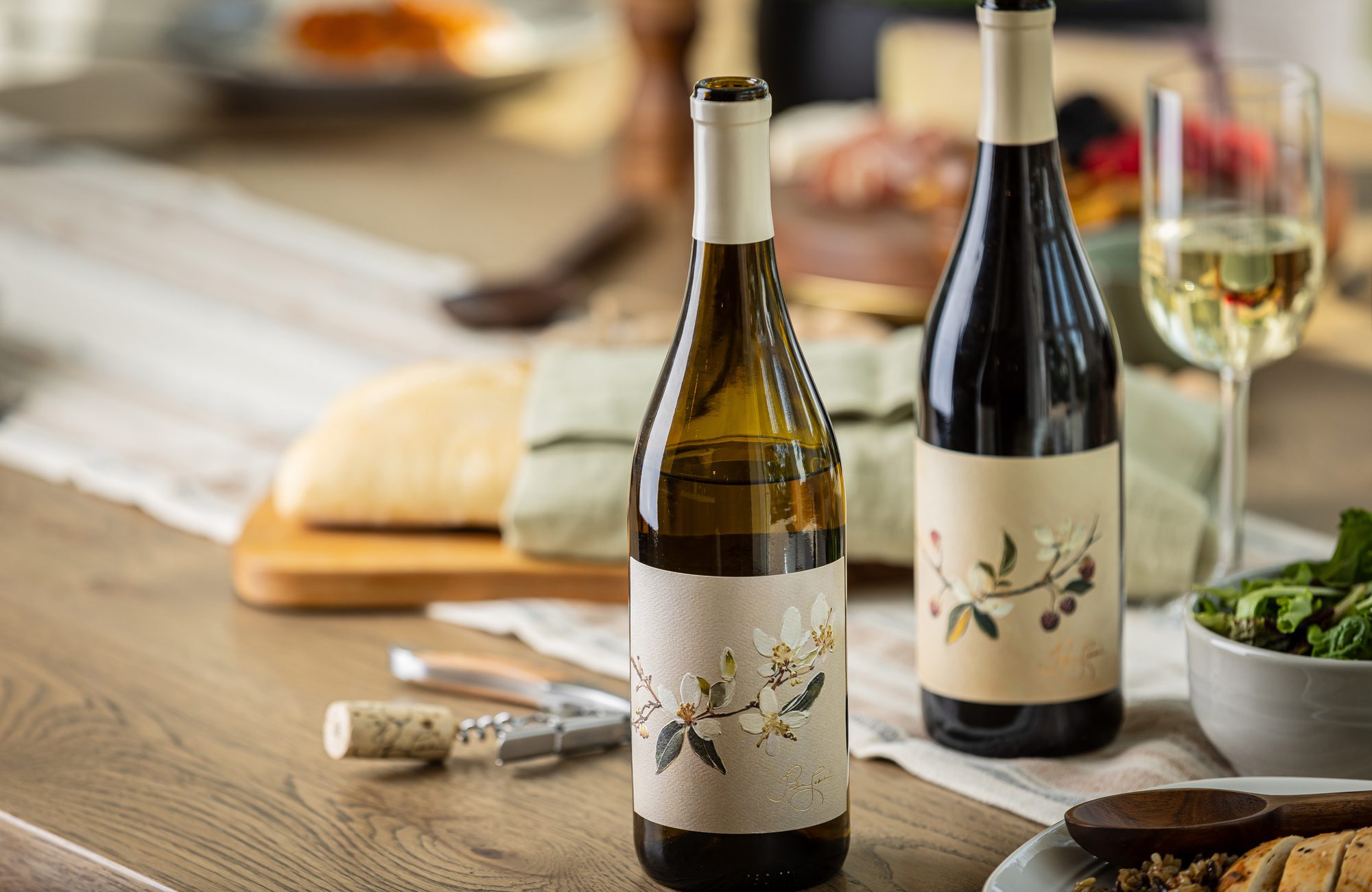 Two floral-labeled wine bottles, a glass of white wine, and a cork with corkscrew on a table with bread and salad