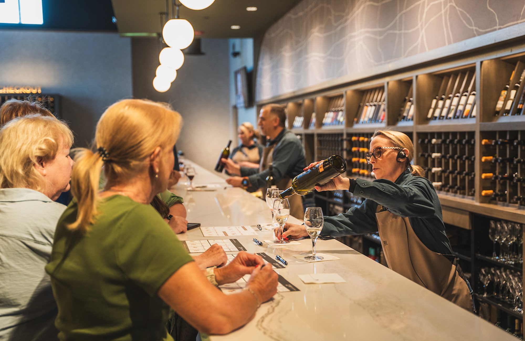 Tasting room staff member pours wine into a glass for customers seated along a long tasting bar.