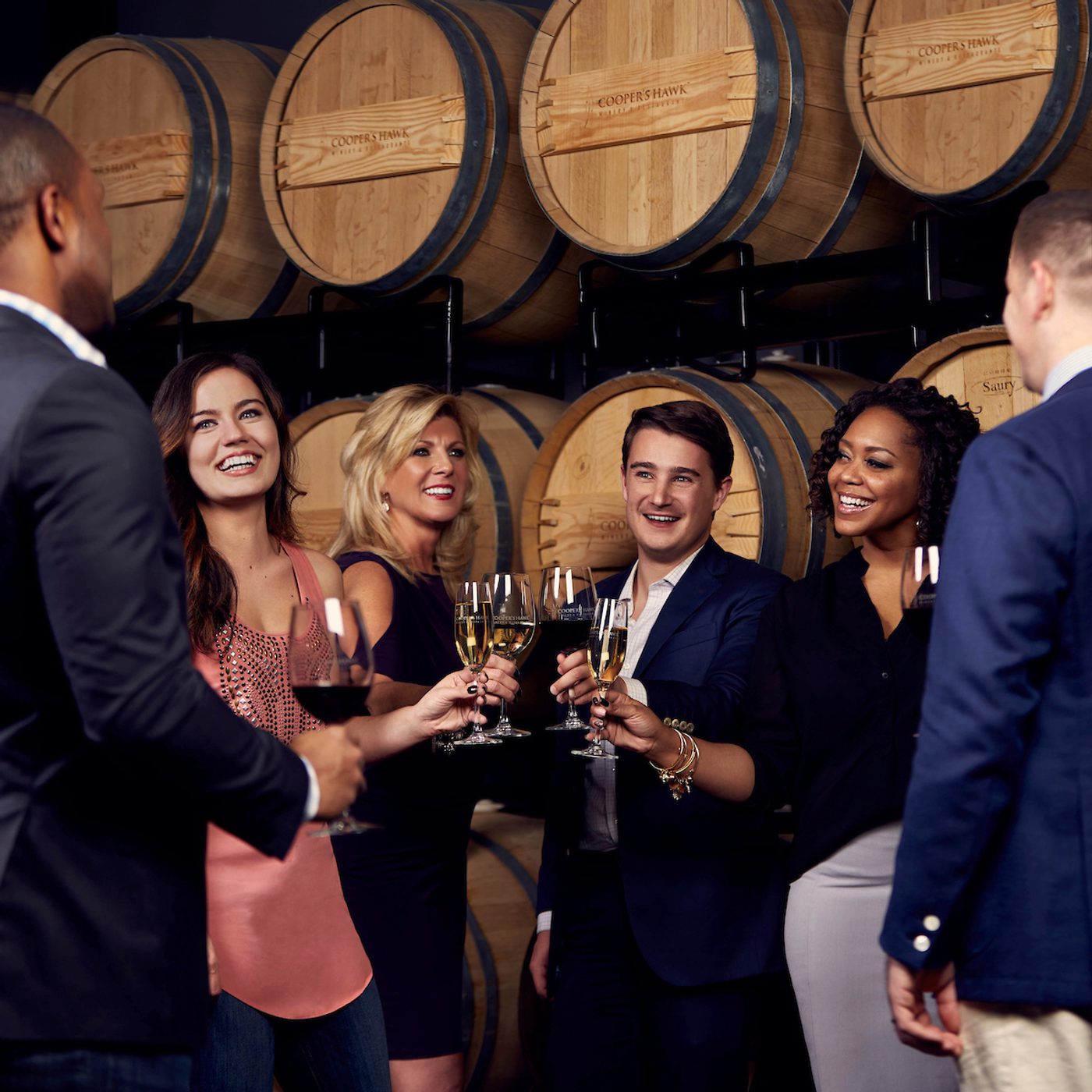 Group of adults toasting with wine glasses in a winery tasting room in front of stacked oak barrels